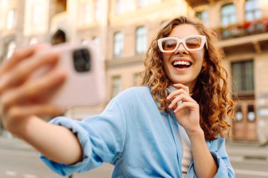 Şık gözlüklü ve modern elbiseli mutlu bir kadın güneşli bir sokakta selfie çekiyor. Yürüme, eğlence, blog yazma, seyahat kavramı