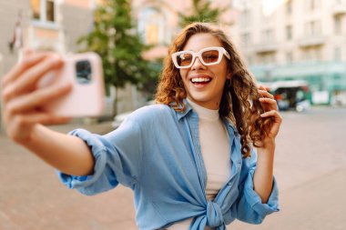 Şık gözlüklü ve modern elbiseli mutlu bir kadın güneşli bir sokakta selfie çekiyor. Yürüme, eğlence, blog yazma, seyahat kavramı