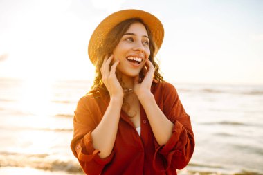 Şapkalı, kollarını havaya kaldırmış, sıcak yaz gününde plaj okyanusunu seyreden mutlu genç bir kadın. Seyahat, hafta sonu, rahatlama ve yaşam tarzı konsepti