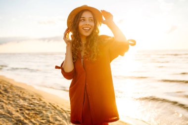 Şapkalı, kollarını havaya kaldırmış, sıcak yaz gününde plaj okyanusunu seyreden mutlu genç bir kadın. Seyahat, hafta sonu, rahatlama ve yaşam tarzı konsepti