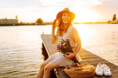 Bir buket yabani çiçekle mutlu genç gün batımında nehirde bir köprüde yürüyor. Düğünün kırsal tarzı.