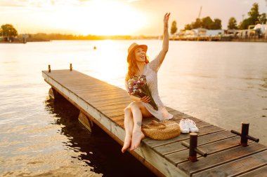 Bir buket yabani çiçekle mutlu genç gün batımında nehirde bir köprüde yürüyor. Düğünün kırsal tarzı.