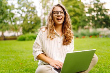 Çimlerin üzerinde oturan genç bir kadın. Yaz parkında uzaktan çalışan bir laptop kullanıyor. Eğitim, iş, blog veya serbest çalışma kavramı