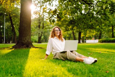Çimlerin üzerinde oturan genç bir kadın. Yaz parkında uzaktan çalışan bir laptop kullanıyor. Eğitim, iş, blog veya serbest çalışma kavramı