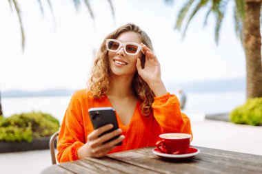 Yazlık kafede kahve içip cep telefonu kullanan mutlu bir kadın. Bloglama, iletişim, seyahat günlüğü