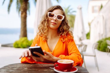 Yazlık kafede kahve içip cep telefonu kullanan mutlu bir kadın. Bloglama, iletişim, seyahat günlüğü
