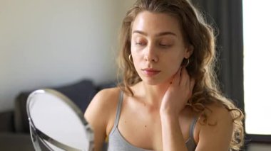 Güzel bir kadın sıcak bir odadaki küçük bir aynadan yansımasına bakar. Genç bir kadın yüz derisine dokunur ve onu muayene eder. Güzellik konsepti, doğallık, cilt bakımı. Yaşam biçimi.