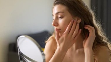 Güzel bir kadın sıcak bir odadaki küçük bir aynadan yansımasına bakar. Genç bir kadın yüz derisine dokunur ve onu muayene eder. Güzellik konsepti, doğallık, cilt bakımı. Yaşam biçimi.