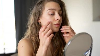 Güzel bir kadın sıcak bir odadaki küçük bir aynadan yansımasına bakar. Genç bir kadın yüz derisine dokunur ve onu muayene eder. Güzellik konsepti, doğallık, cilt bakımı. Yaşam biçimi.