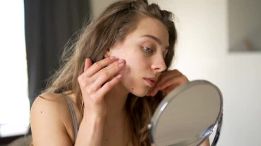 Güzel bir kadın sıcak bir odadaki küçük bir aynadan yansımasına bakar. Genç bir kadın yüz derisine dokunur ve onu muayene eder. Güzellik konsepti, doğallık, cilt bakımı. Yaşam biçimi.