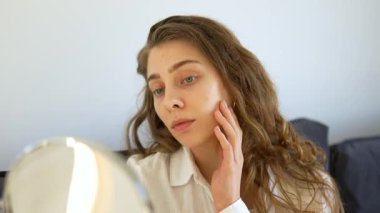 Güzel bir kadın sıcak bir odadaki küçük bir aynadan yansımasına bakar. Genç bir kadın yüz derisine dokunur ve onu muayene eder. Güzellik konsepti, doğallık, cilt bakımı. Yaşam biçimi.