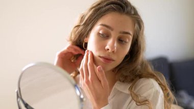 Güzel bir kadın sıcak bir odadaki küçük bir aynadan yansımasına bakar. Genç bir kadın yüz derisine dokunur ve onu muayene eder. Güzellik konsepti, doğallık, cilt bakımı. Yaşam biçimi.