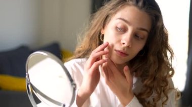 Güzel bir kadın sıcak bir odadaki küçük bir aynadan yansımasına bakar. Genç bir kadın yüz derisine dokunur ve onu muayene eder. Güzellik konsepti, doğallık, cilt bakımı. Yaşam biçimi.