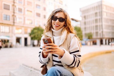 Şık bir kadın güneşli bir şehirde telefonla dolaşıyor. Güneş gözlüklü güzel bir turist gün batımında blog çekiyor. Turizm konsepti, yürüyüşler. Etkin yaşam biçimi.