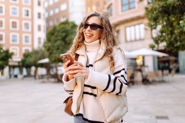 Şık bir kadın güneşli bir şehirde telefonla dolaşıyor. Güneş gözlüklü güzel bir turist gün batımında blog çekiyor. Turizm konsepti, yürüyüşler. Etkin yaşam biçimi.