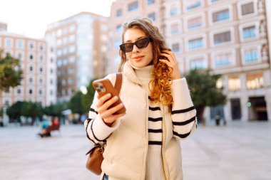 Şık bir kadın güneşli bir şehirde telefonla dolaşıyor. Güneş gözlüklü güzel bir turist gün batımında blog çekiyor. Turizm konsepti, yürüyüşler. Etkin yaşam biçimi.