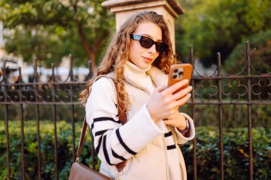 Şık bir kadın güneşli bir şehirde telefonla dolaşıyor. Güneş gözlüklü güzel bir turist gün batımında blog çekiyor. Turizm konsepti, yürüyüşler. Etkin yaşam biçimi.