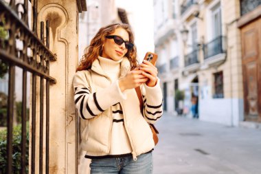 Şık bir kadın güneşli bir şehirde telefonla dolaşıyor. Güneş gözlüklü güzel bir turist gün batımında blog çekiyor. Turizm konsepti, yürüyüşler. Etkin yaşam biçimi.