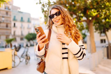 Şık bir kadın güneşli bir şehirde telefonla dolaşıyor. Güneş gözlüklü güzel bir turist gün batımında blog çekiyor. Turizm konsepti, yürüyüşler. Etkin yaşam biçimi.