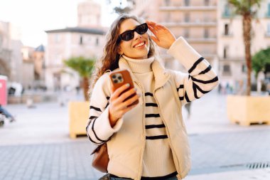 Şık bir kadın güneşli bir şehirde telefonla dolaşıyor. Güneş gözlüklü güzel bir turist gün batımında blog çekiyor. Turizm konsepti, yürüyüşler. Etkin yaşam biçimi.