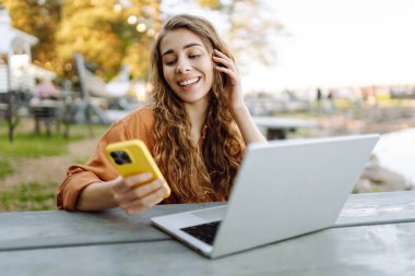 Parkta dizüstü bilgisayarla çalışan telefonlu gülümseyen bir kadın. Güzel bayan serbest yazar havanın tadını çıkarıyor ve dışarıda çalışıyor. Öğrenme, blog yazma kavramı.