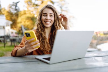 Parkta dizüstü bilgisayarla çalışan telefonlu gülümseyen bir kadın. Güzel bayan serbest yazar havanın tadını çıkarıyor ve dışarıda çalışıyor. Öğrenme, blog yazma kavramı.