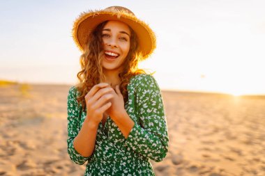 Parlak bir elbise ve hasır şapkalı mutlu bir kadının portresi plajda eğleniyor. Güzel bir kadın sahilde günbatımının tadını çıkarıyor. Rahatlama kavramı, güzellik.