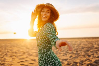 Parlak bir elbise ve hasır şapkalı mutlu bir kadının portresi plajda eğleniyor. Güzel bir kadın sahilde günbatımının tadını çıkarıyor. Rahatlama kavramı, güzellik.