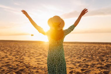 Parlak bir elbise ve hasır şapkalı mutlu bir kadının portresi plajda eğleniyor. Güzel bir kadın sahilde günbatımının tadını çıkarıyor. Rahatlama kavramı, güzellik.