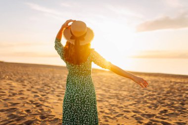 Parlak bir elbise ve hasır şapkalı mutlu bir kadının portresi plajda eğleniyor. Güzel bir kadın sahilde günbatımının tadını çıkarıyor. Rahatlama kavramı, güzellik.