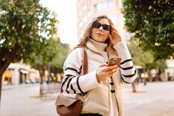 Şık bir kadın güneşli bir şehirde telefonla dolaşıyor. Güneş gözlüklü güzel bir turist gün batımında blog çekiyor. Turizm konsepti, yürüyüşler. Etkin yaşam biçimi.