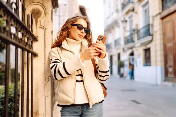 Şık bir kadın güneşli bir şehirde telefonla dolaşıyor. Güneş gözlüklü güzel bir turist gün batımında blog çekiyor. Turizm konsepti, yürüyüşler. Etkin yaşam biçimi.