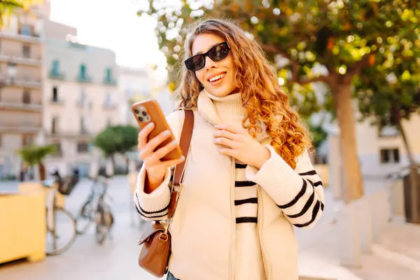 Şık bir kadın güneşli bir şehirde telefonla dolaşıyor. Güneş gözlüklü güzel bir turist gün batımında blog çekiyor. Turizm konsepti, yürüyüşler. Etkin yaşam biçimi.