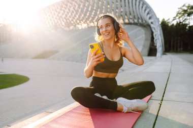 Spor kıyafetli bir kadın paspasın üzerinde egzersiz yapıyor ve gün batımında müzik dinliyor. Modern bir parkta kablosuz kulaklık takan sportif bir kadın. Spor konsepti. Etkin yaşam biçimi.