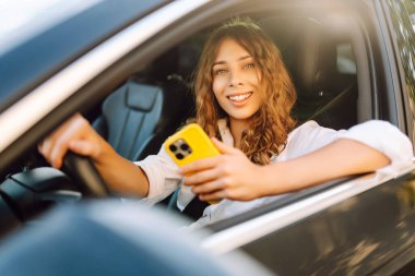Kıvırcık saçlı, elinde telefonla araba kullanan genç bir kadın. Elinde telefonla mutlu bir kadın sürücü güneşli bir günün tadını çıkarıyor. Teknoloji, navigasyon ve ulaşım kavramı.