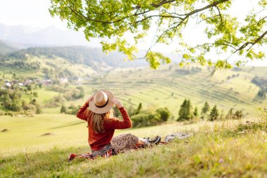 Şapkalı güzel kadın gün batımında Yeşil Tepe 'den manzaranın tadını çıkarıyor. Genç bayan gezgin rahatlar ve manzara karşısında özgürlüğünü hisseder. Seyahat, eğlence, yürüyüş konsepti.