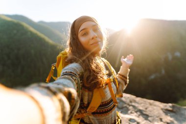 Sarı bir sırt çantasıyla günbatımı dağ arka planında selfie çeken genç bir bayan yürüyüşçü. Telefonu olan güzel bir kadın uçurumun kenarında özgürce eğleniyor. Doğa konsepti.