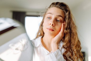 Küçük bir aynada cildini inceleyen genç bir kadının portresi. Güzel bir kadın evde eğleniyor. Güzellik ve cilt bakımı.