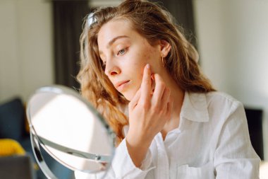 Küçük bir aynada cildini inceleyen genç bir kadının portresi. Güzel bir kadın evde eğleniyor. Güzellik ve cilt bakımı.