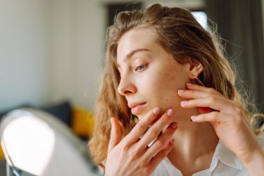 Küçük bir aynada cildini inceleyen genç bir kadının portresi. Güzel bir kadın evde eğleniyor. Güzellik ve cilt bakımı.