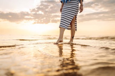 Gün batımında deniz kenarında yürüyen güzel kadın bacaklarının yakın çekimi. Deniz kıyısında yürüyen ve manzaranın tadını çıkaran bir kadın. Boş zaman konsepti. Etkin yaşam biçimi.