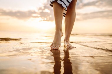 Gün batımında deniz kenarında yürüyen güzel kadın bacaklarının yakın çekimi. Deniz kıyısında yürüyen ve manzaranın tadını çıkaran bir kadın. Boş zaman konsepti. Etkin yaşam biçimi.