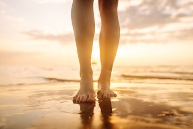 Gün batımında deniz kenarında yürüyen güzel kadın bacaklarının yakın çekimi. Deniz kıyısında yürüyen ve manzaranın tadını çıkaran bir kadın. Boş zaman konsepti. Etkin yaşam biçimi.