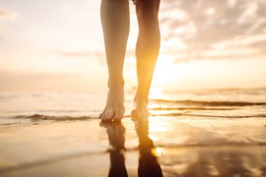Gün batımında deniz kenarında yürüyen güzel kadın bacaklarının yakın çekimi. Deniz kıyısında yürüyen ve manzaranın tadını çıkaran bir kadın. Boş zaman konsepti. Etkin yaşam biçimi.