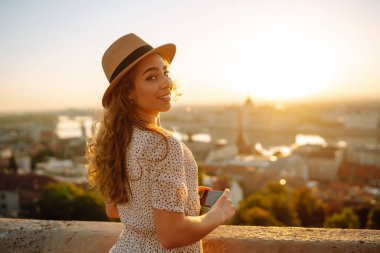 Telefonla şapkalı neşeli kadın günbatımı manzarasının keyfini çıkarıyor. Güzel bir turist fotoğraf çekiyor ya da dışarıda blog yazıyor. Turizm, blog konsepti.
