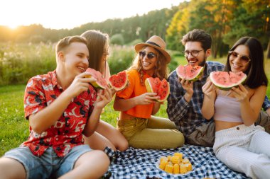 Mutlu arkadaşlar piknikte karpuz yiyorlar. Genç kadınlar ve erkekler gün batımında birlikte dışarıda vakit geçiriyorlar. Arkadaşlık kavramı, eğlence..
