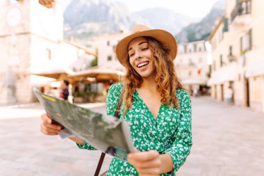 Parlak elbiseli gülümseyen kadın şehrin haritasına bakıyor. Turist, antik bir kentin mimarinin arka planına kıyasla navigasyonunu araştırıyor. Seyahat, tatil konsepti.