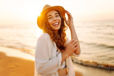 Güneş batarken sahilde eğlenen şapkalı genç bir kadının portresi. Güzel bayan turist özgürlüğü hissediyor ve dışarıda vakit geçirmeyi seviyor. Yaşam tarzı, eğlence..