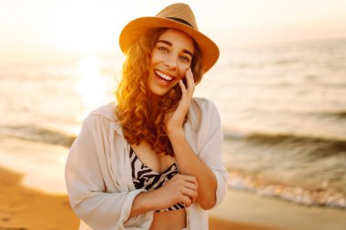 Güneş batarken sahilde eğlenen şapkalı genç bir kadının portresi. Güzel bayan turist özgürlüğü hissediyor ve dışarıda vakit geçirmeyi seviyor. Yaşam tarzı, eğlence..