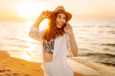 Güneş batarken sahilde eğlenen şapkalı genç bir kadının portresi. Güzel bayan turist özgürlüğü hissediyor ve dışarıda vakit geçirmeyi seviyor. Yaşam tarzı, eğlence..